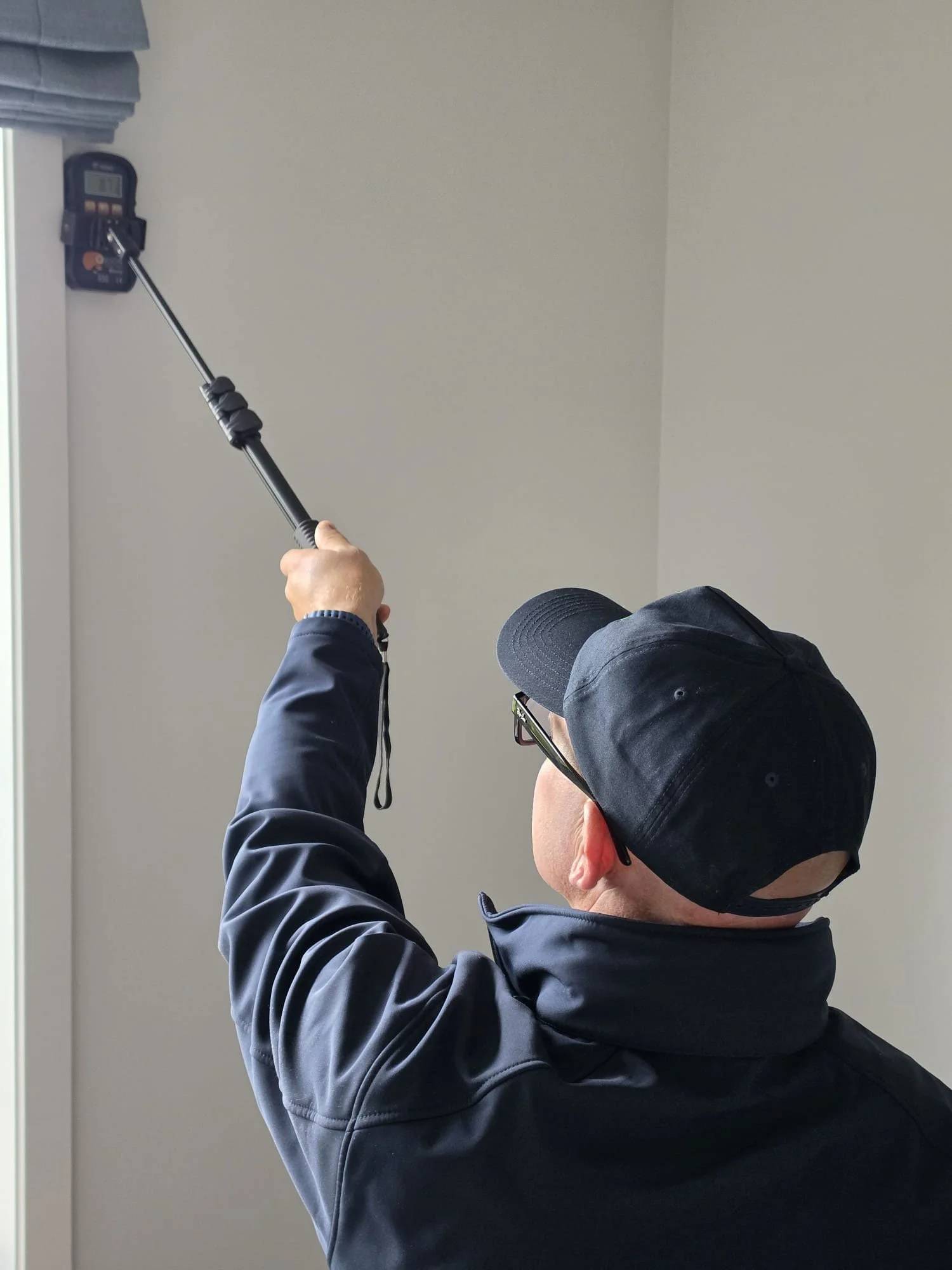 A person wearing a black cap, glasses, and a dark jacket is using a grabber tool to reach an object near the ceiling.