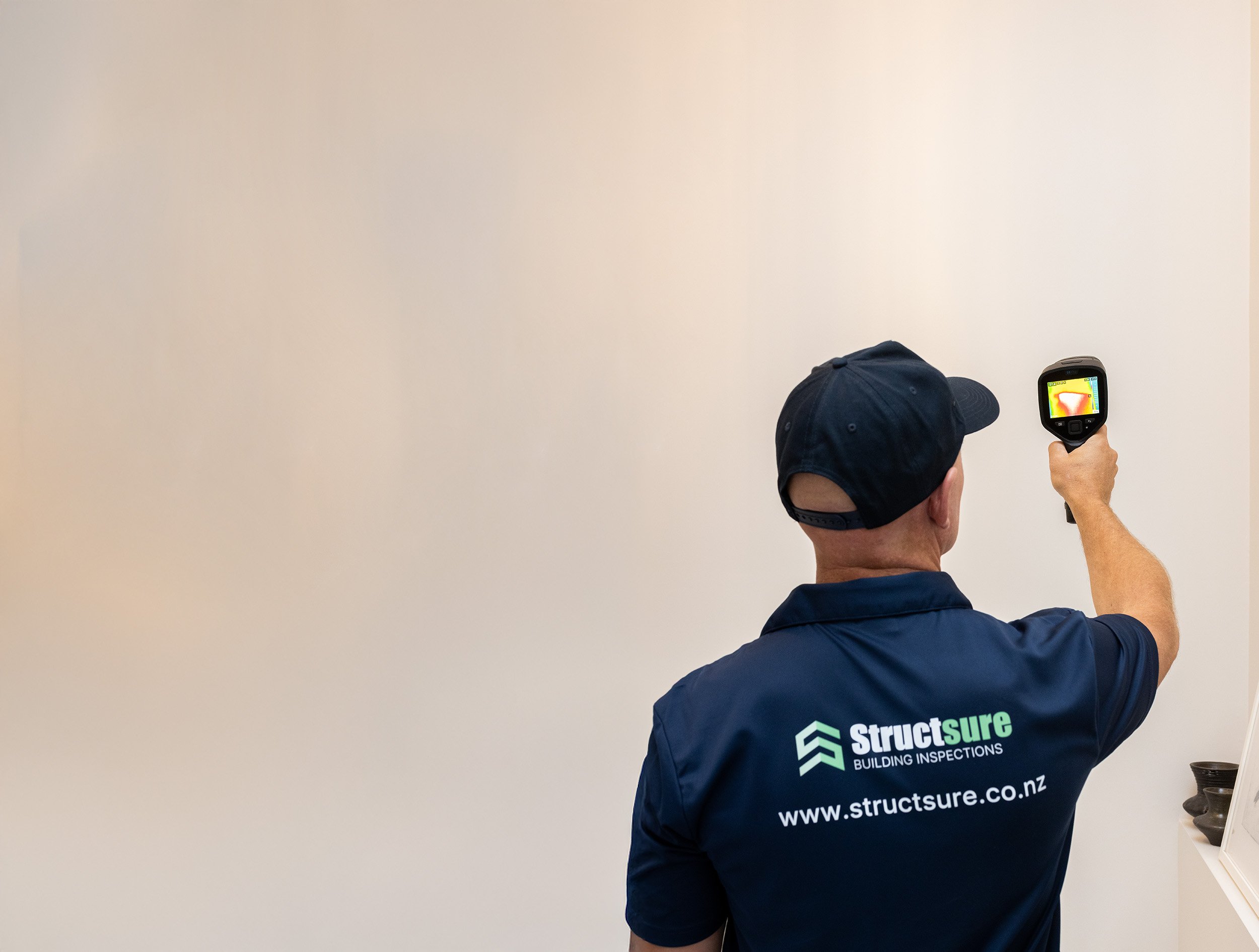 A person wearing a navy blue shirt and a baseball cap is checking the interior of a wall with a thermal imaging camera. The back of the person's shirt displays the logo and website of Structsure Building Inspections.