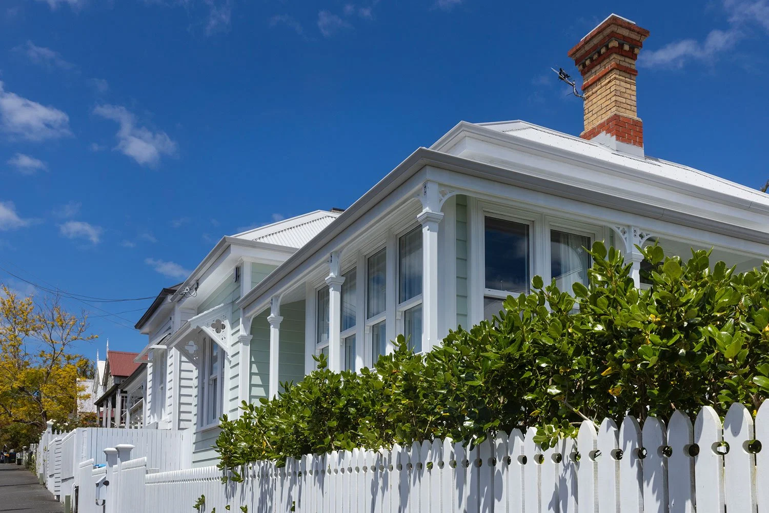 White Victorian house with a metal roof, large windows, a brick chimney, lush green bushes and a white picket fence, under a clear blue sky.