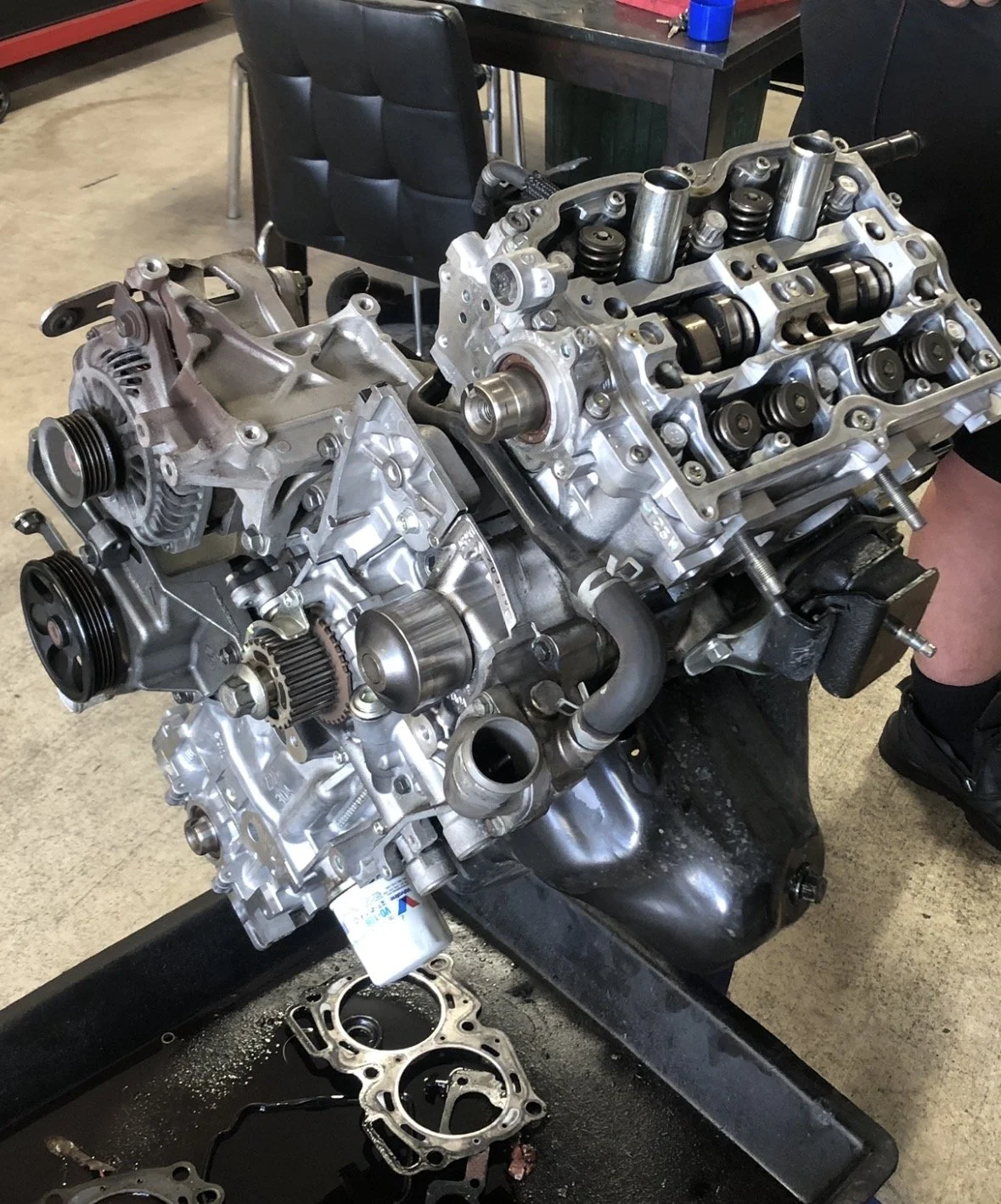An automotive engine on a workbench with disassembled parts and gaskets nearby, in a garage or workshop setting.