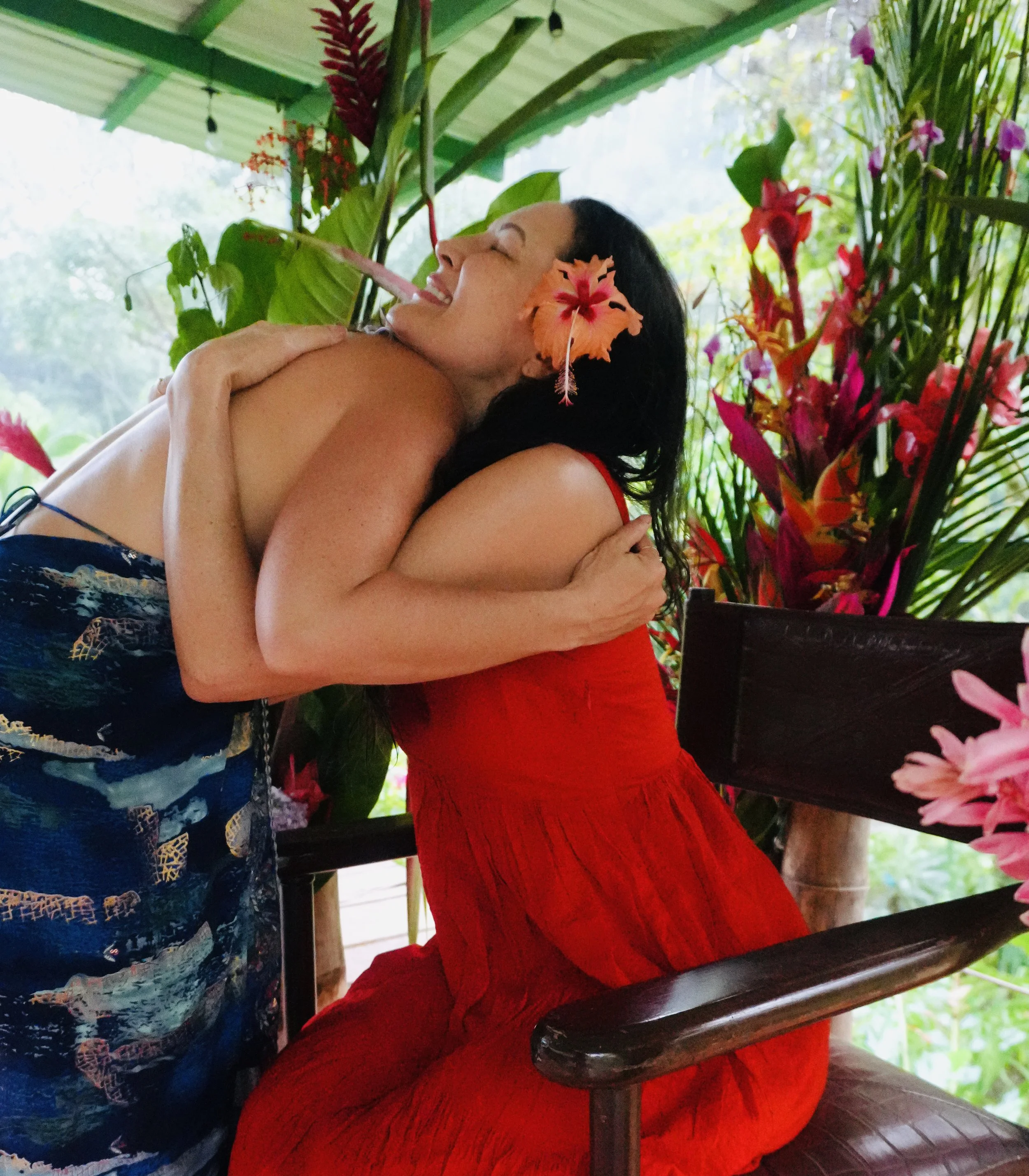 Two women hugging each other in a tropical setting with lush green plants and vibrant flowers, one wearing a red dress and the other in a patterned top, smiling and enjoying a joyful moment.