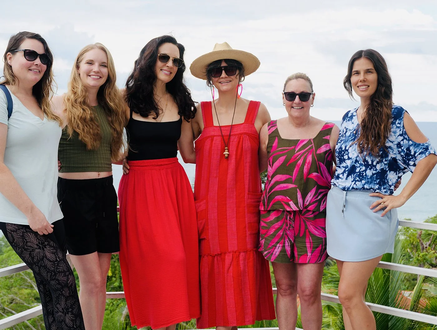 Seven women standing together outdoors, with a cloudy sky and trees in the background, smiling and posing for the photo.