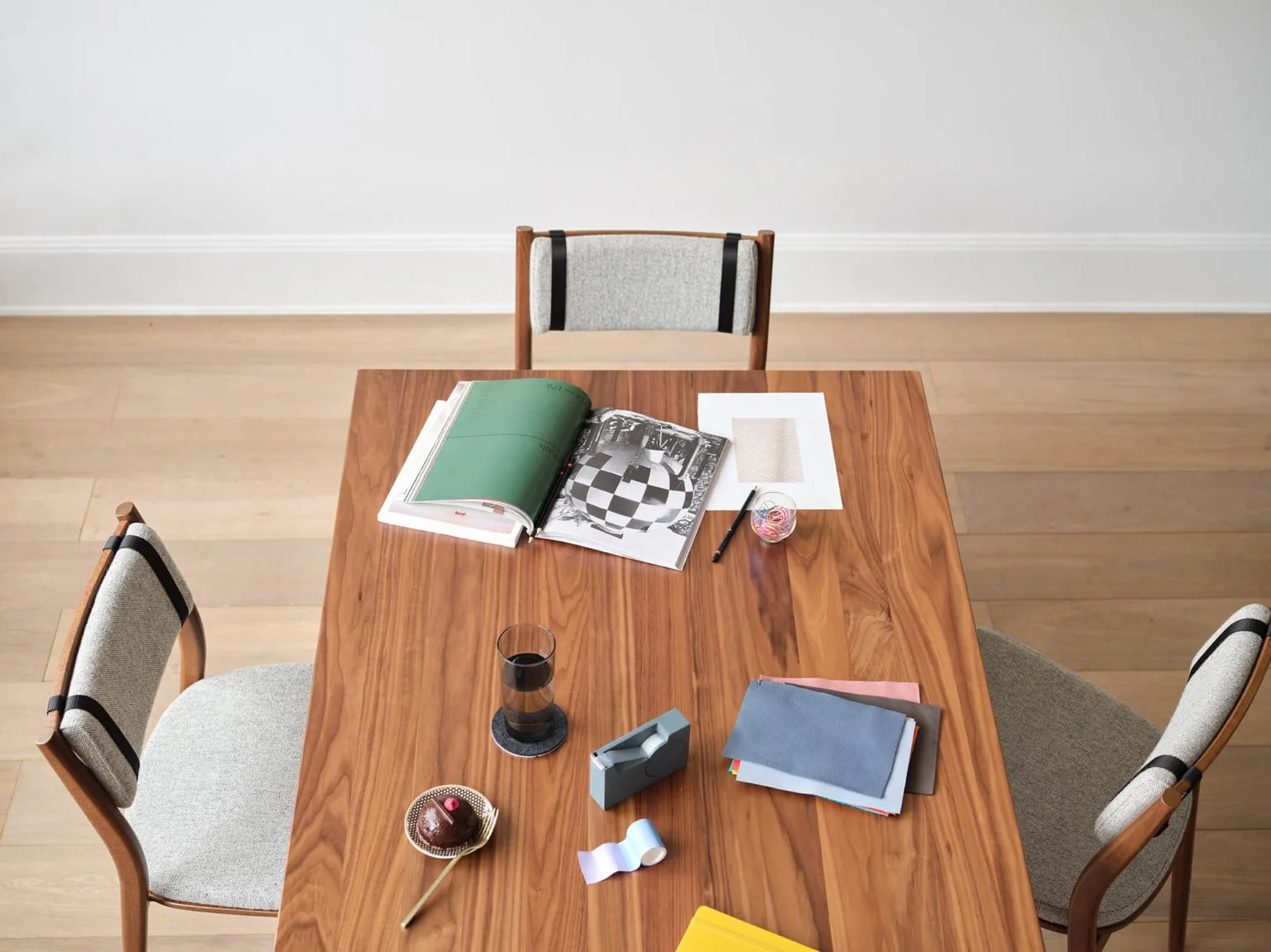 Top-down view of a wooden dining table with three chairs, various books, papers, a glass of a dark beverage, a cupcake with a pink candle, a tape dispenser, paper scraps, and writing utensils on it.