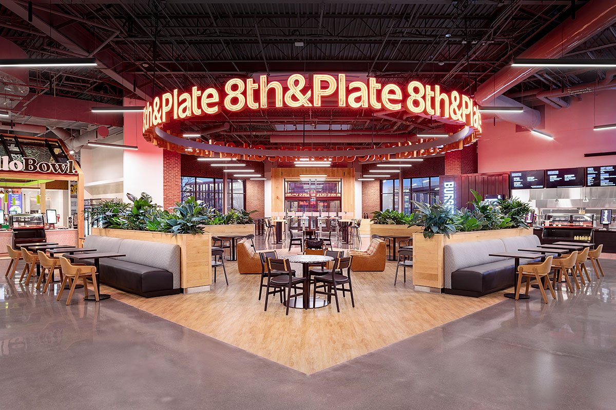 Interior of a modern food court area with seating, plants, and a sign for 8th & Plate restaurant.