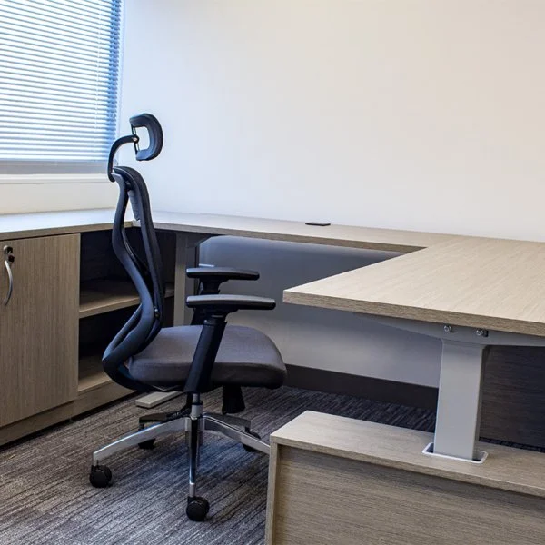 Empty office corner with a black ergonomic chair and a large L-shaped wooden desk in front of a window with closed blinds.