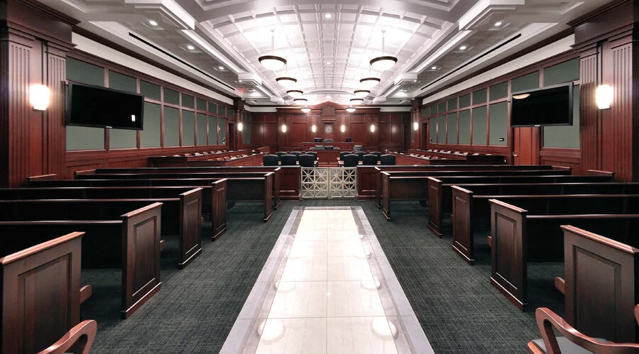 Empty courtroom with wooden benches, two large monitors, and a judge's bench at the front.