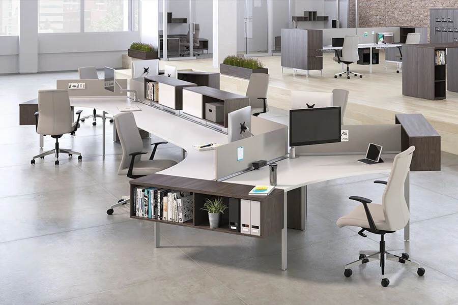 Modern open office space with white and wooden desks, computer monitors, office chairs, and shelves with books and plants.