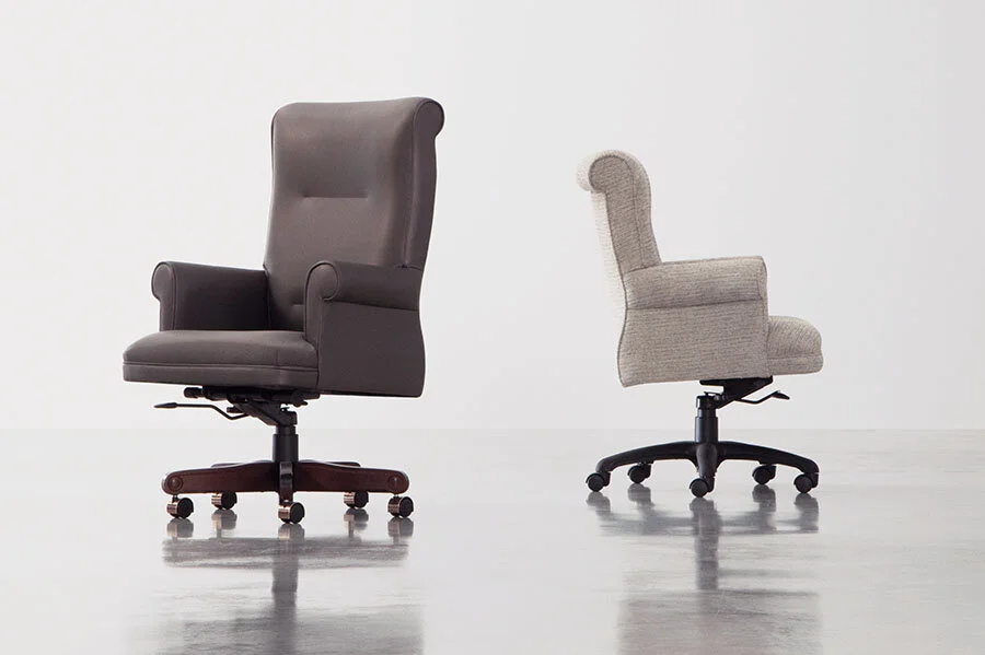 Two office chairs, one in front of the other, on a reflective floor against a plain background. The first chair is dark gray leather with a high back and armrests, and the second chair is light gray fabric with a lower back and no armrests.