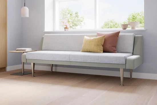 A light gray modern sofa with three pillows in front of a window with white frame, and a small side table with books next to it.