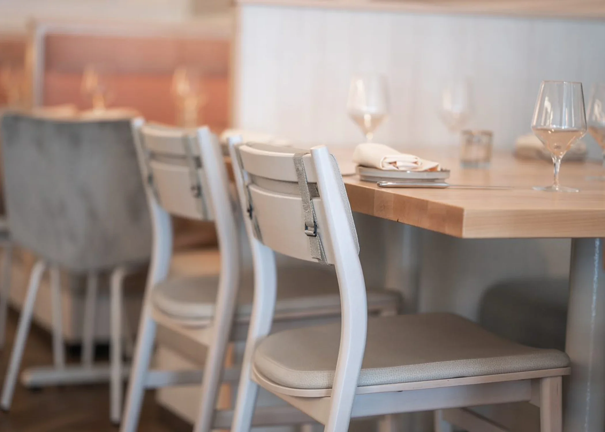 Empty dining table set with three wine glasses, a folded napkin, and cutlery in a restaurant with white chairs and a wooden wall in the background.