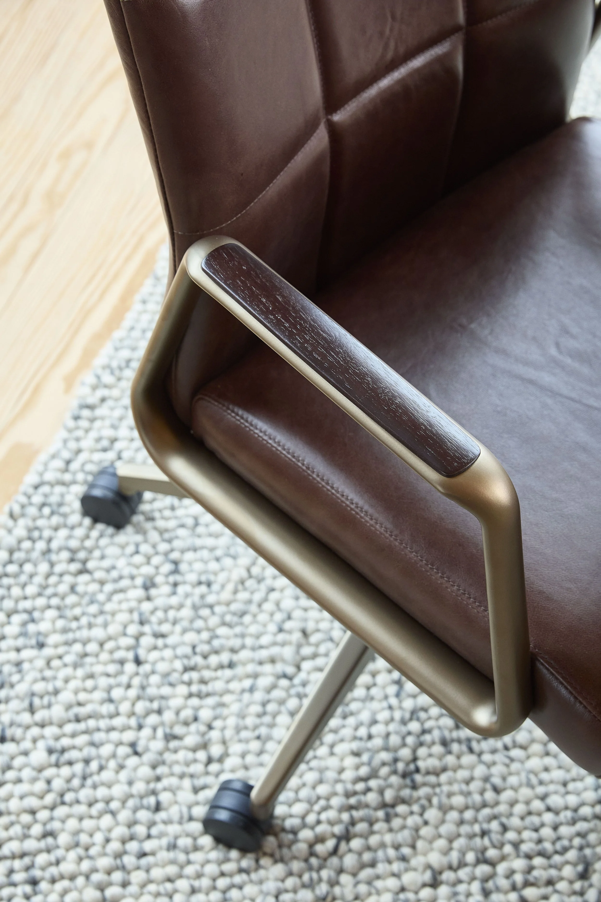 Close-up of a brown leather office chair with a metal armrest on a beige textured rug, with wooden floor visible in the background.