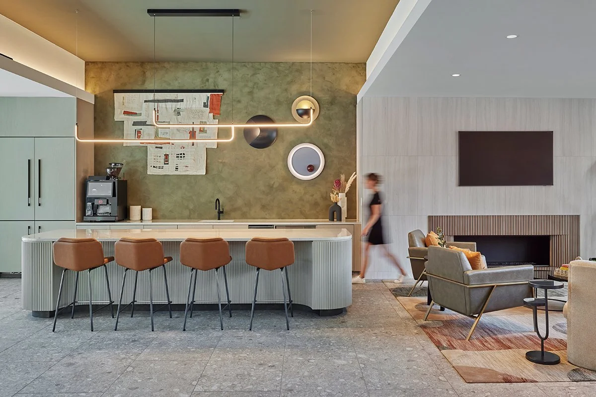 Modern kitchen and lounge area with a green wall, barstools, and a woman walking past in the background.