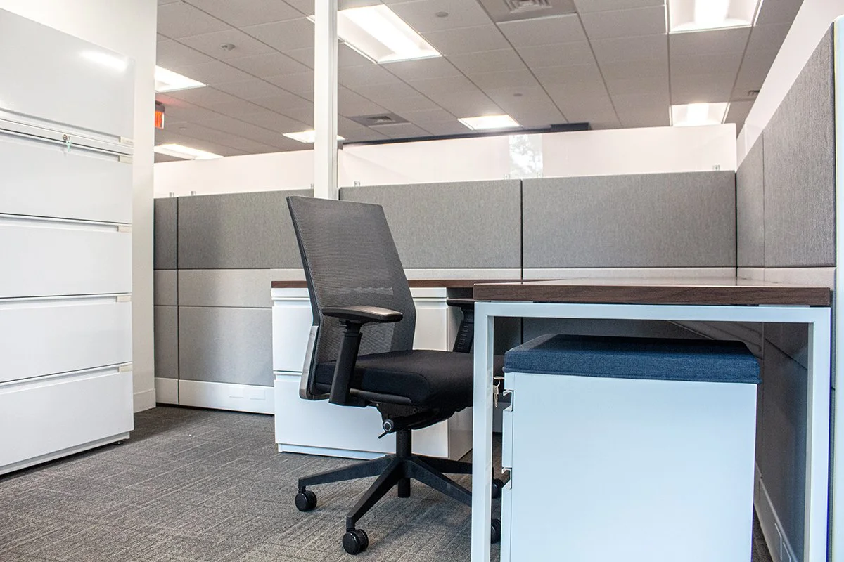 Empty office cubicle with a black mesh office chair and a white desk with a dark wood top.