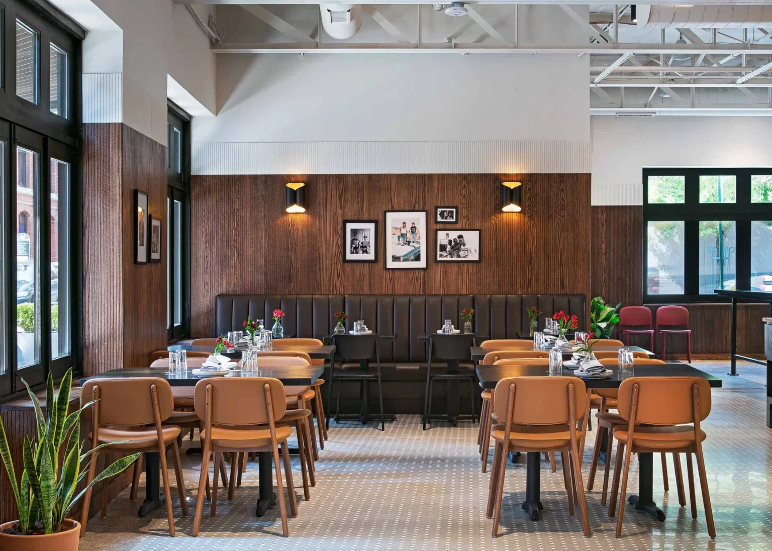 Empty restaurant dining area with tables set with glasses and flowers, brown chairs, wooden wall decor, black framed photos, large windows, and some red chairs in the background.