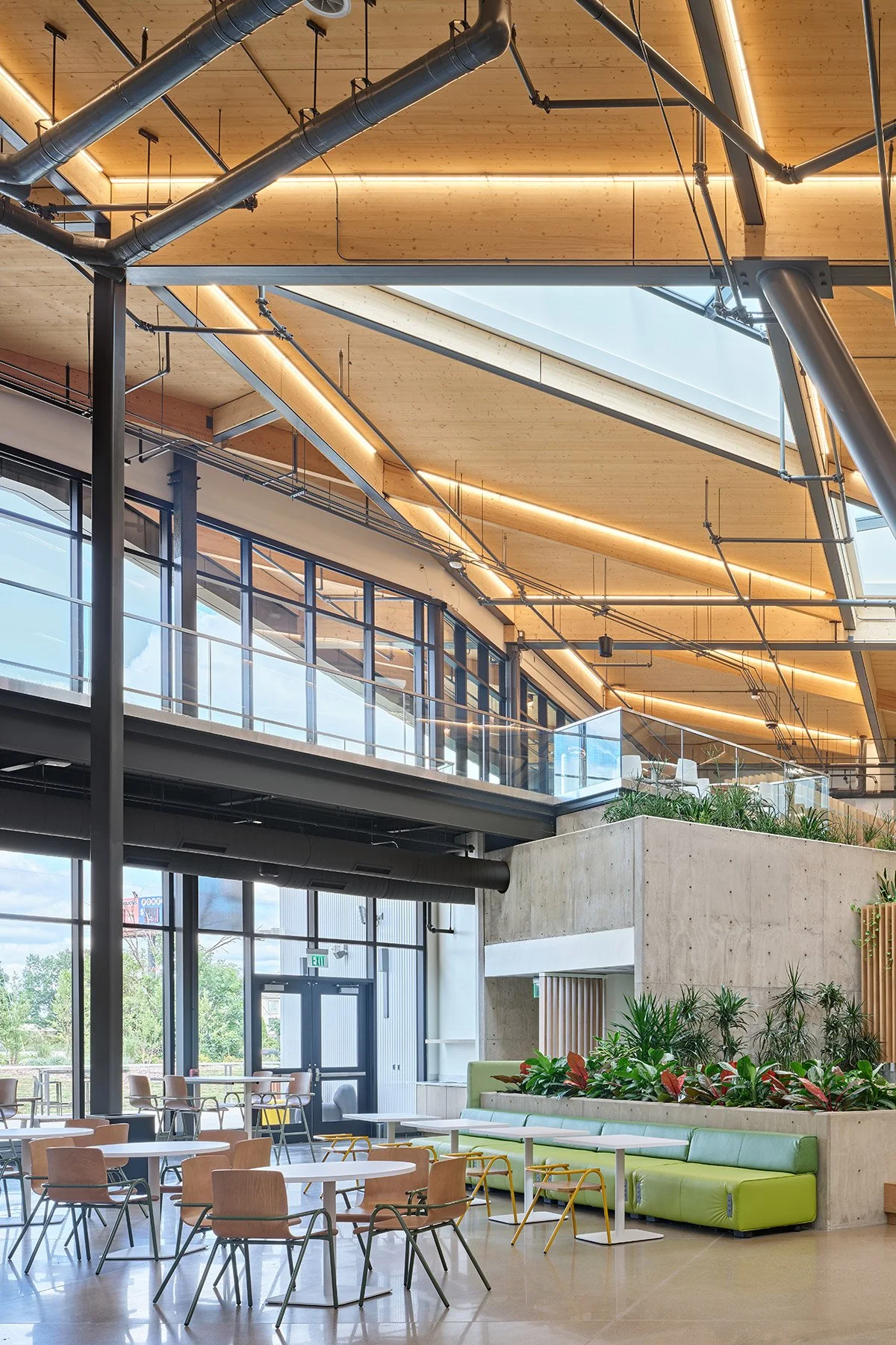 Interior of a modern, spacious building with large glass windows, wooden and concrete elements, and exposed ductwork, featuring seating areas with tables and chairs, and green plants.
