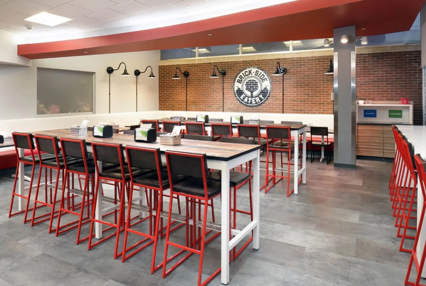 Modern restaurant interior with high tables, red and black chairs, brick wall, and a sign reading 'Brick Side Eatery' in the background.