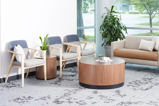 A waiting area with four gray-cushioned chairs with wooden armrests, a round wooden coffee table, and a beige sofa with white pillows, near large windows with greenery outside.