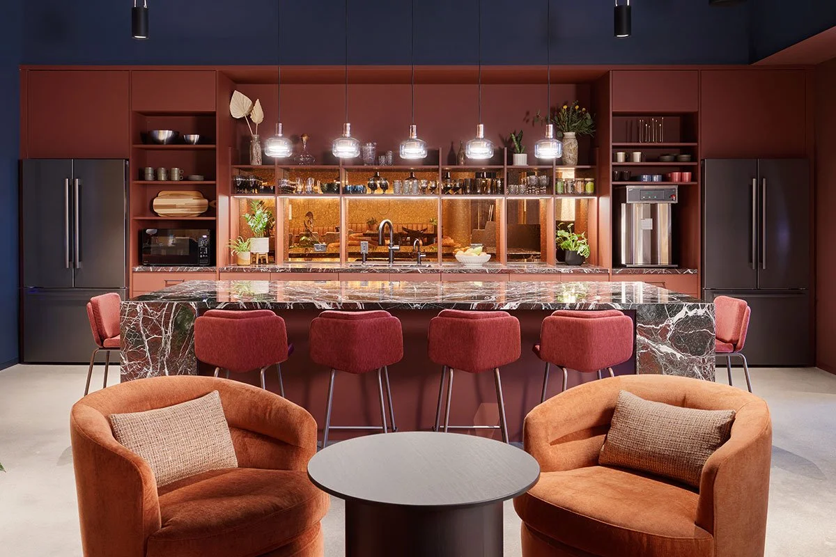 Interior of a modern kitchen with a large bar counter made of dark marble, surrounded by six red upholstered bar stools. There are two orange armchairs with cushions and a small round table in the seating area in the foreground. The background features dark red cabinetry, open shelving with dishes and plants, a microwave, and a coffee machine, with pendant lighting hanging above the bar.