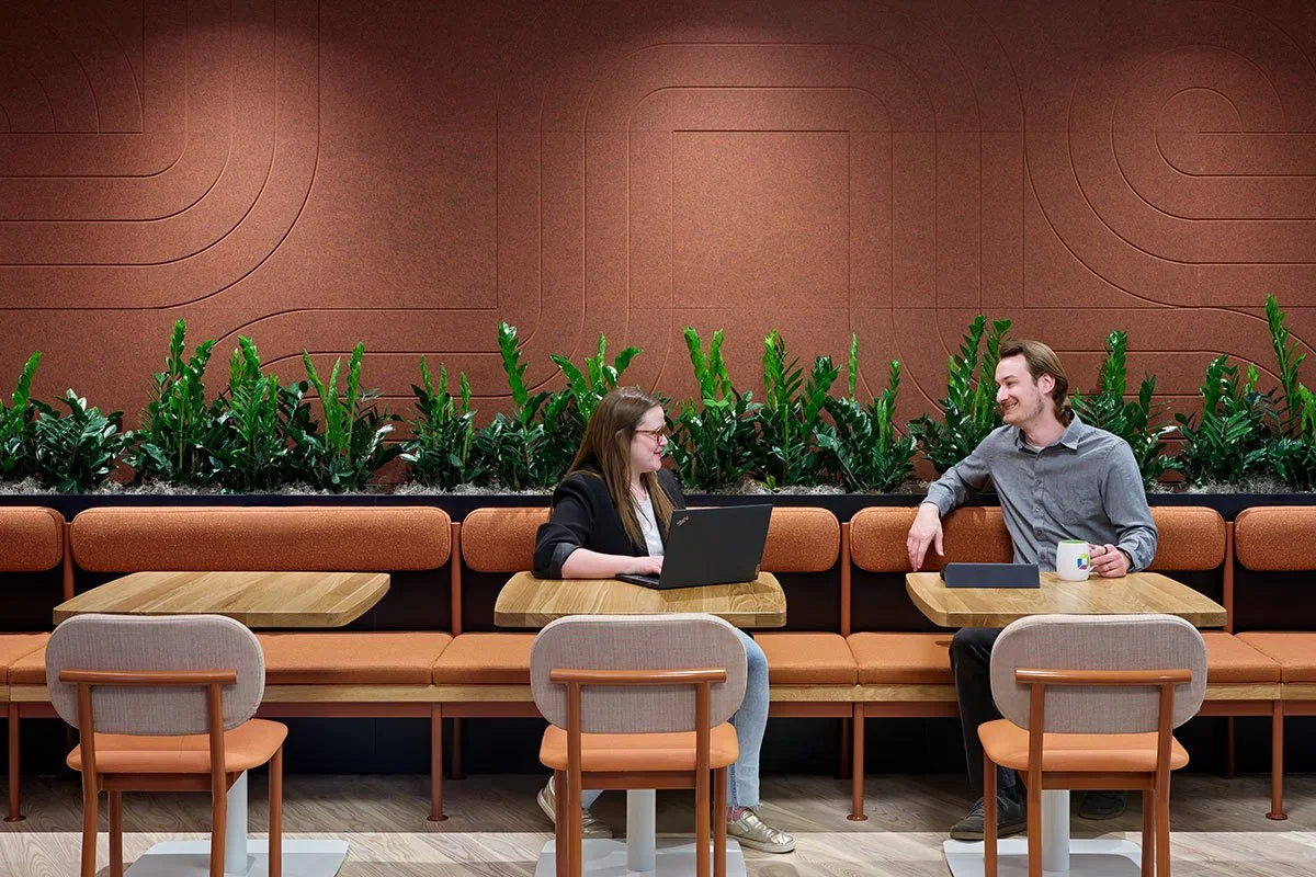 Two people sitting at a booth in a modern, stylish cafe or lounge, engaging in conversation. The woman has a laptop open in front of her, and the man is holding a coffee mug, smiling. Behind them, there is a decorative wall with geometric patterns and green plants.
