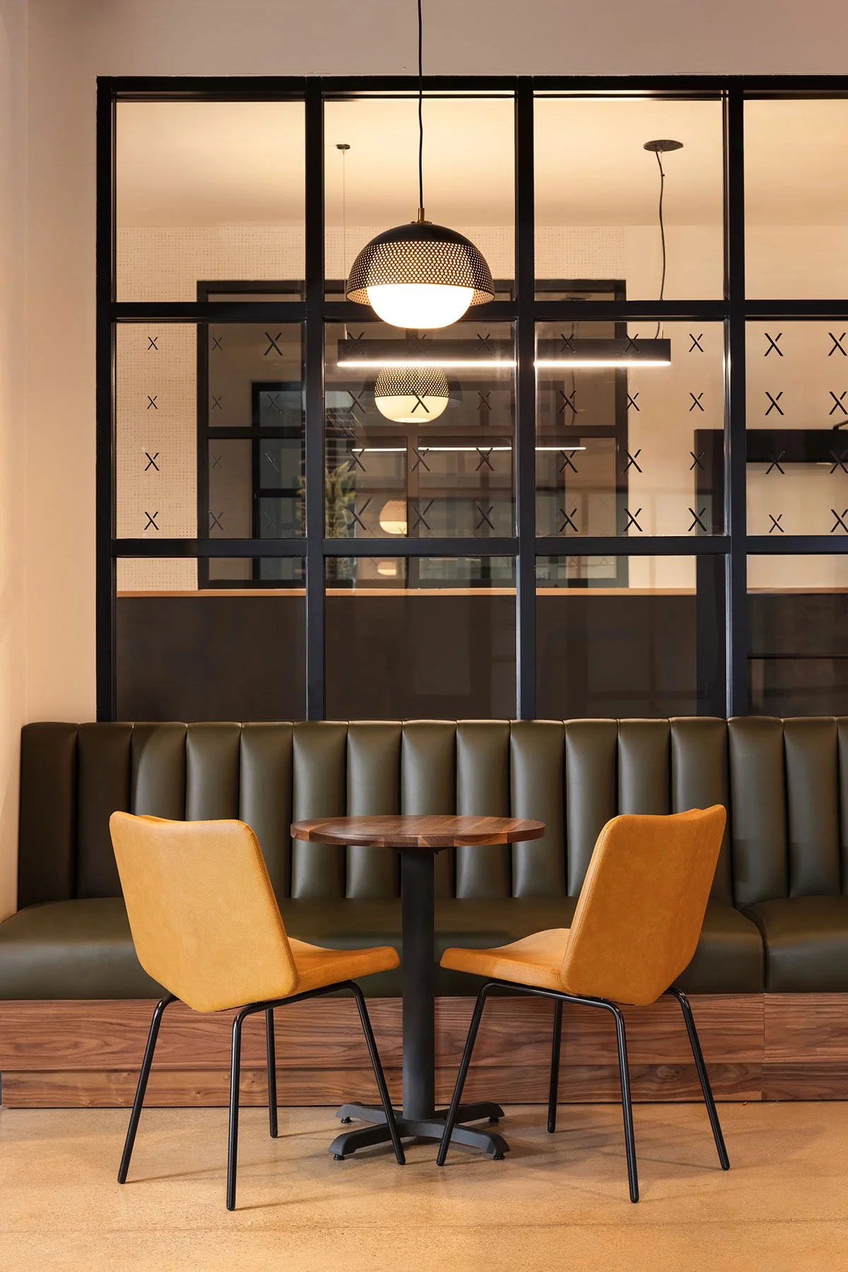 Interior of a modern coffee shop with a small round table, two mustard-colored chairs, a green leather bench, decorative black metal grid wall, and hanging ceiling lights.