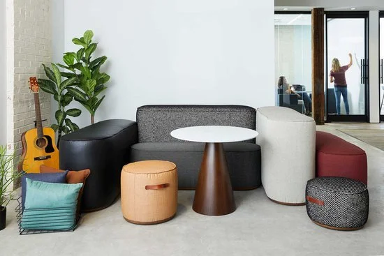 Modern living room with a gray sofa, assorted colorful cushions, a small round table, an indoor plant, a guitar, and a woman taking a photo through a glass door.