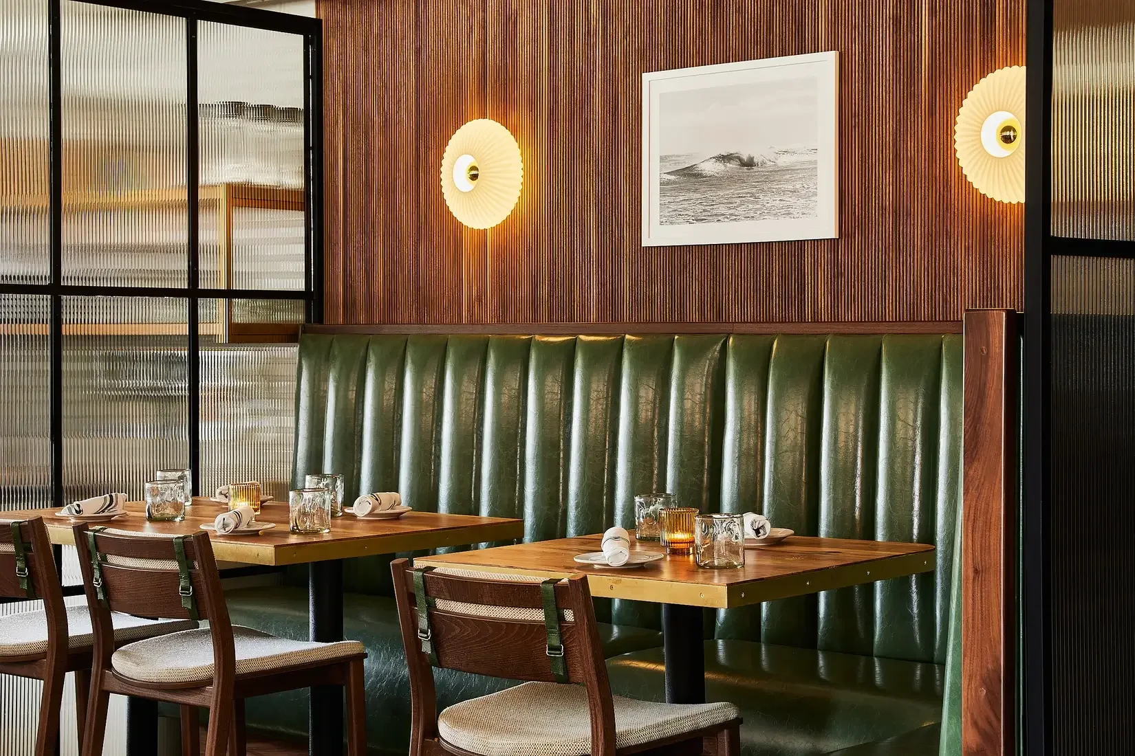 Interior of a restaurant with wooden walls, a green leather banquette, two wooden tables set with glasses and rolled napkins, and modern wall sconces.