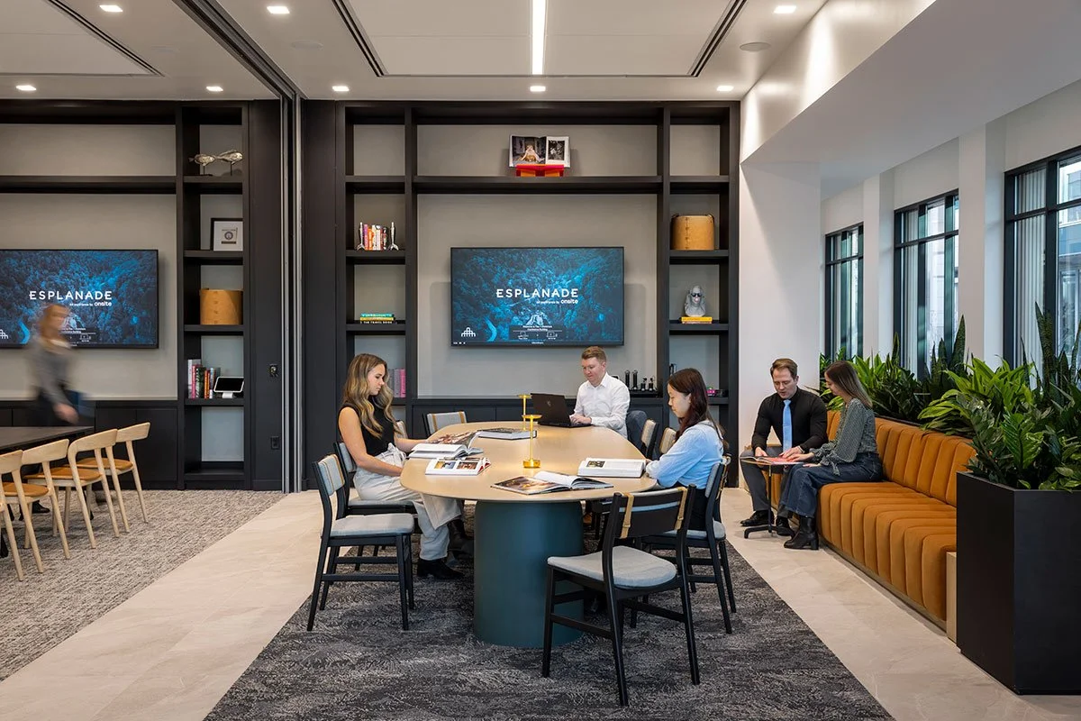 Modern conference room with five people seated around an oval table, looking at books and laptops. The room features ample natural light, built-in shelves behind with a TV screen displaying 'ESPLANADE,' and large windows on the right. There’s a mustard-colored bench with two people sitting, and indoor plants on the right.