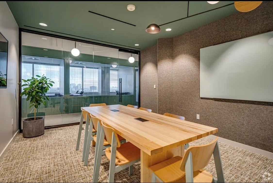 Modern conference room with a wooden table, yellow chairs, a large whiteboard, a potted plant, and a glass wall with a view of an office space and cityscape outside.