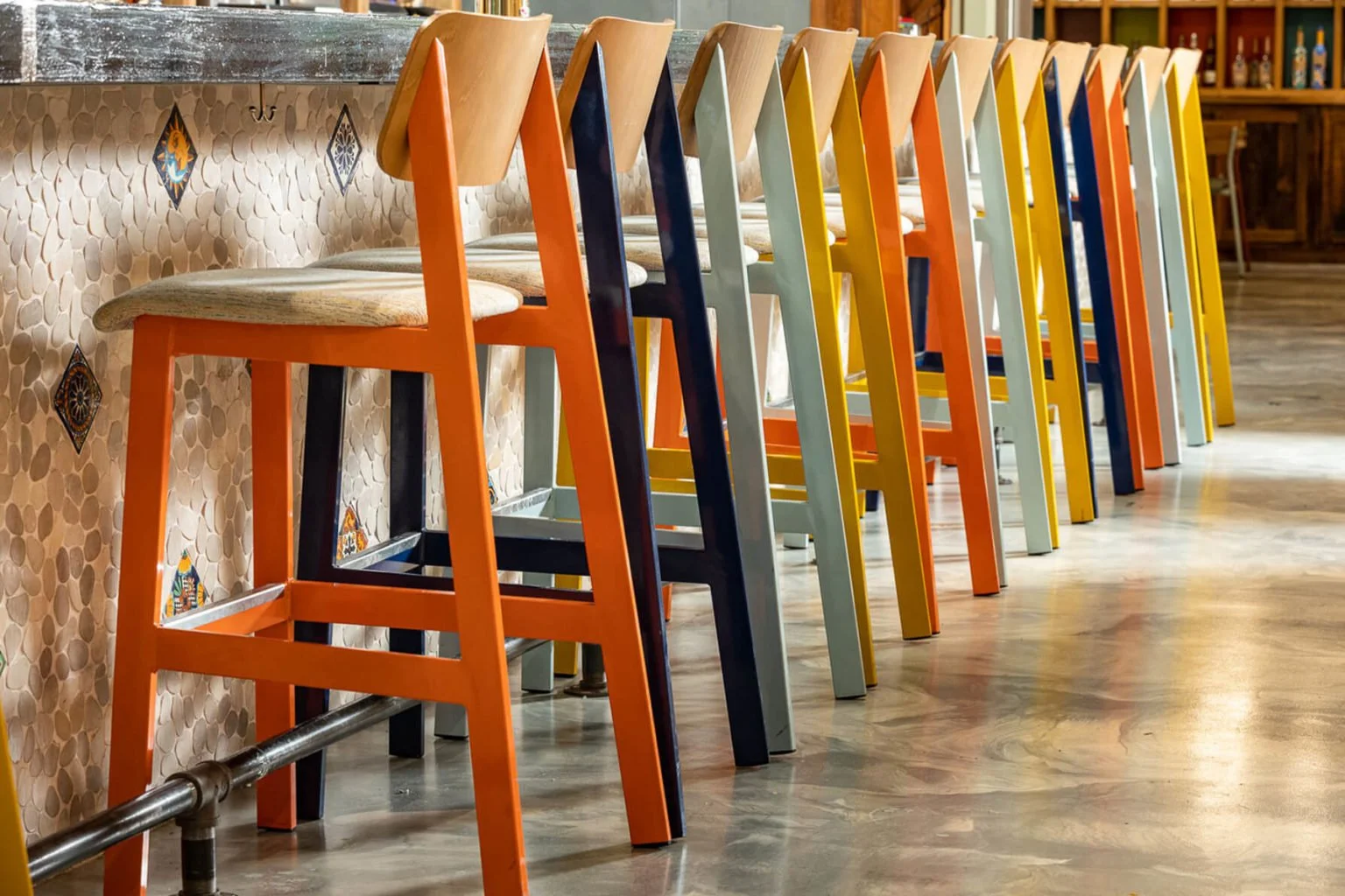 A row of colorful bar stools with wooden seats and different painted legs aligned along a bar counter in a restaurant or cafe. The background shows a tiled wall and part of a wooden cabinet.