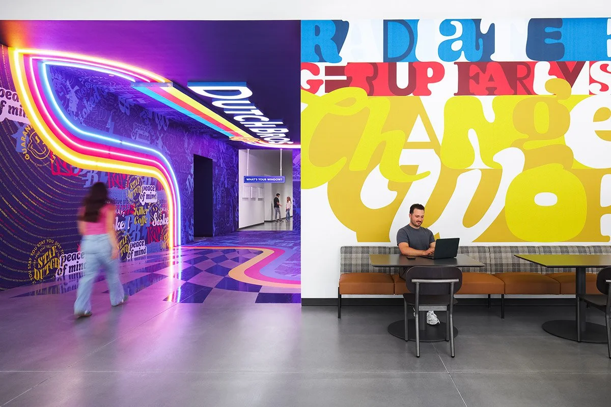 Interior of a colorful, modern space with neon lighting, vibrant wall art, and a man working on a laptop at a table.