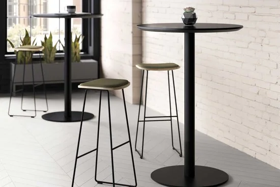 Two high-top black tables with beige stools in a modern, industrial-style room with large windows, white brick walls, and white wooden floors.