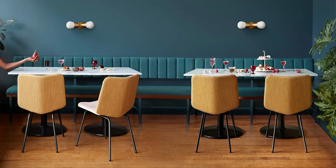 A dining room with two tables set for a celebration, decorated with desserts, berries, and drinks, and a person holding a strawberry on the left side.