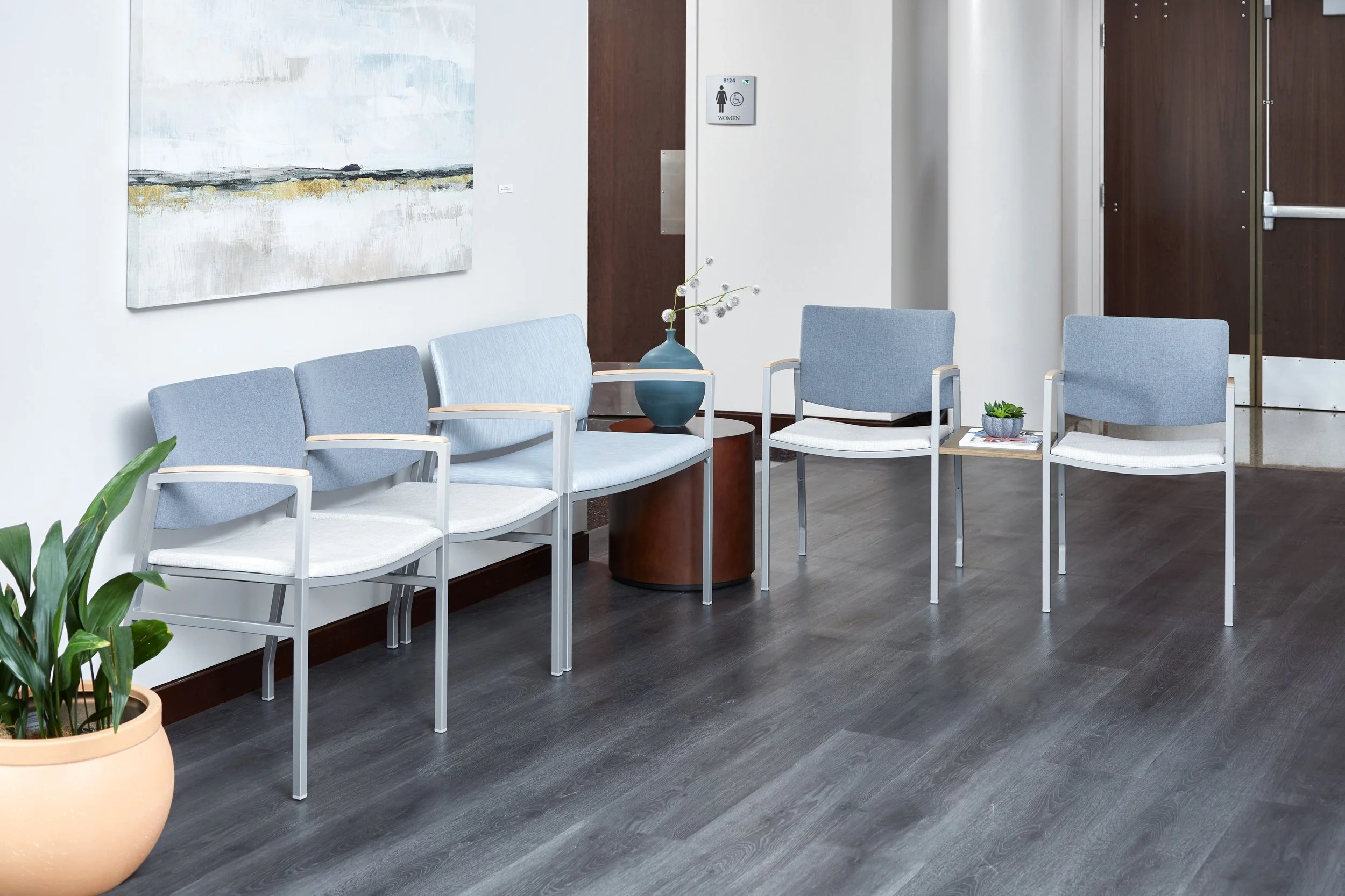 Waiting area with four light blue chairs, a potted plant, a small round table with a vase, and a wall decorated with abstract art in a medical office or clinic.