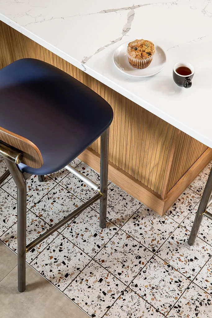 A close-up view of a white marble countertop with a muffin on a white plate and a cup of coffee. Part of a wooden-paneled counter and a black chair with metal legs are also visible.