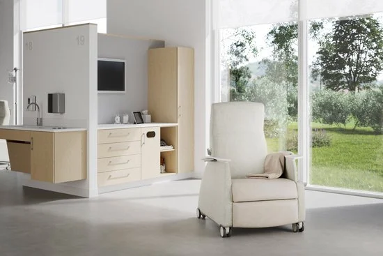 Hospital room with a white armchair, large window showing trees and grass, and medical equipment in the background.