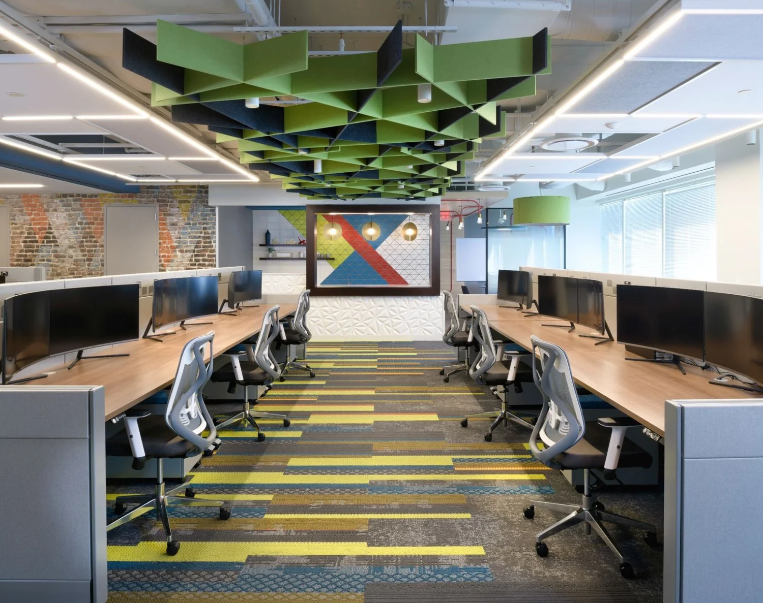 Modern office conference room with computers, ergonomic chairs, brick wall, artistic ceiling design, and colorful patterned carpet.