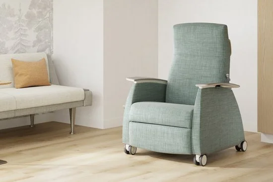 A mint green reclining lift chair with armrests on wheels, next to a cream-colored sofa with two pillows, in a modern living room with light wood flooring and neutral walls.