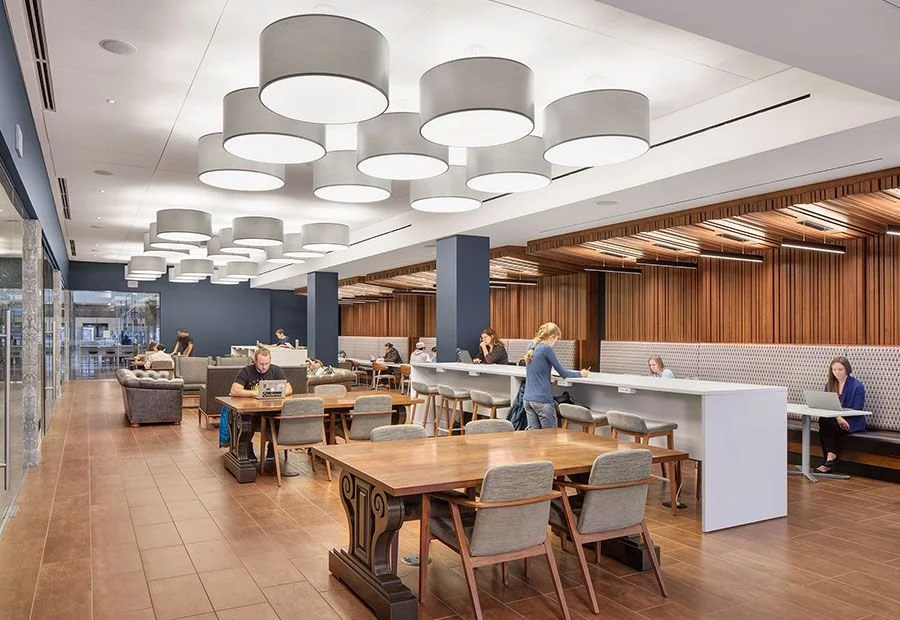 Coffee shop or cafe with patrons working and socializing, modern decor, large pendant lights, wooden accents, seating areas with tables and chairs, and a large window.