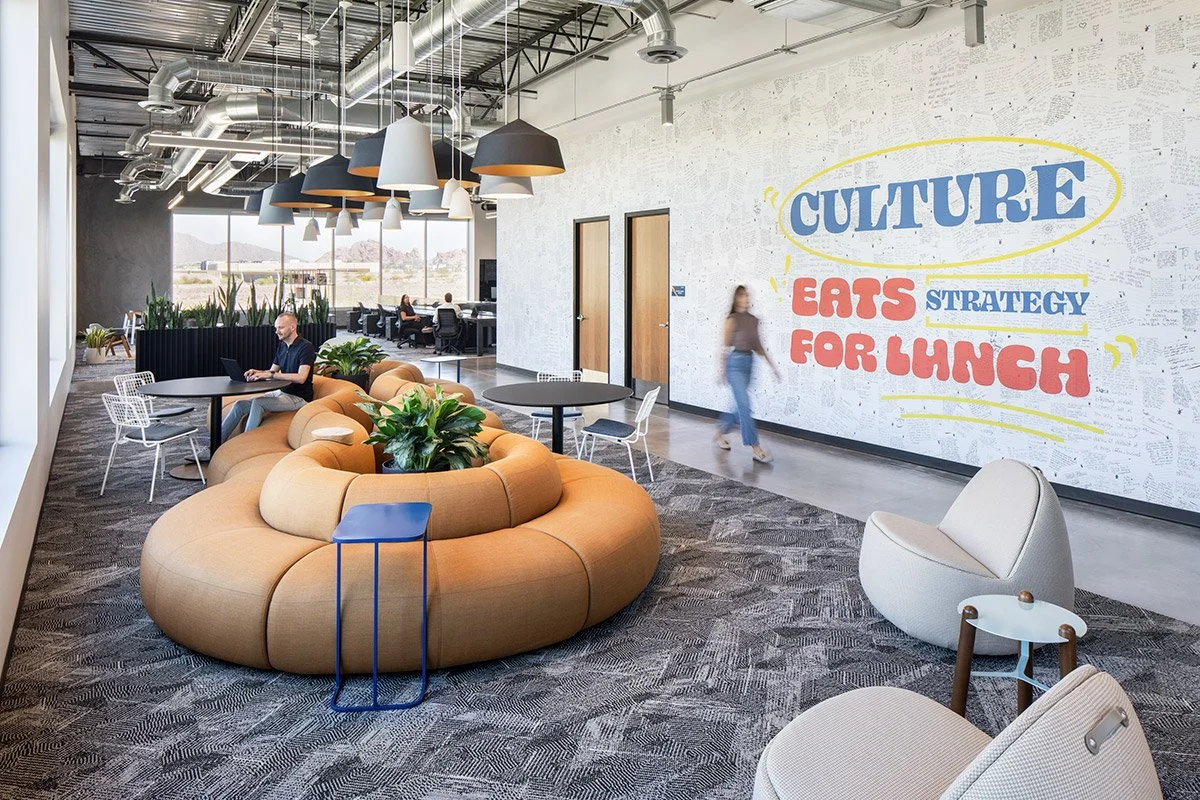 Modern office lounge with round seating, plants, tables, and a large wall mural reading 'Culture Eats Strategy for Lunch' in bold colors.