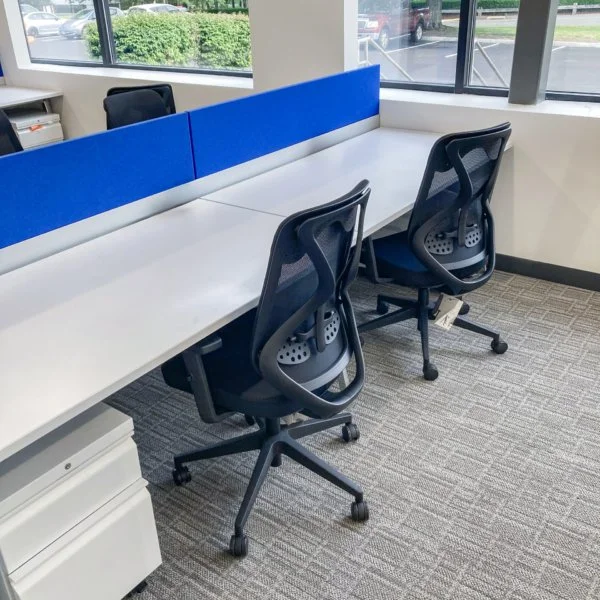 Office cubicle with white desk, black ergonomic chairs, blue privacy panels, and large windows showing a parking lot outside.
