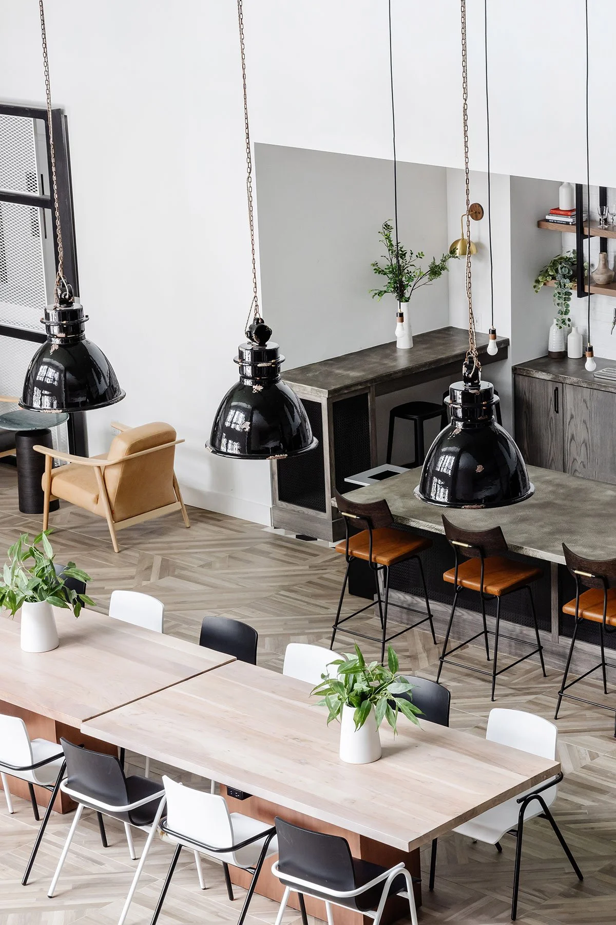 Modern dining area with a large wooden table, black and white chairs, potted plants, pendant lights, and a kitchen counter with bar stools and minimalist decor.