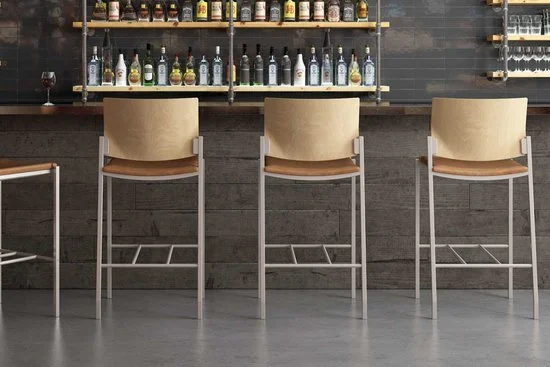 Bar counter with bottles and glasses, four beige bar stools in front, dark tiled wall in background.