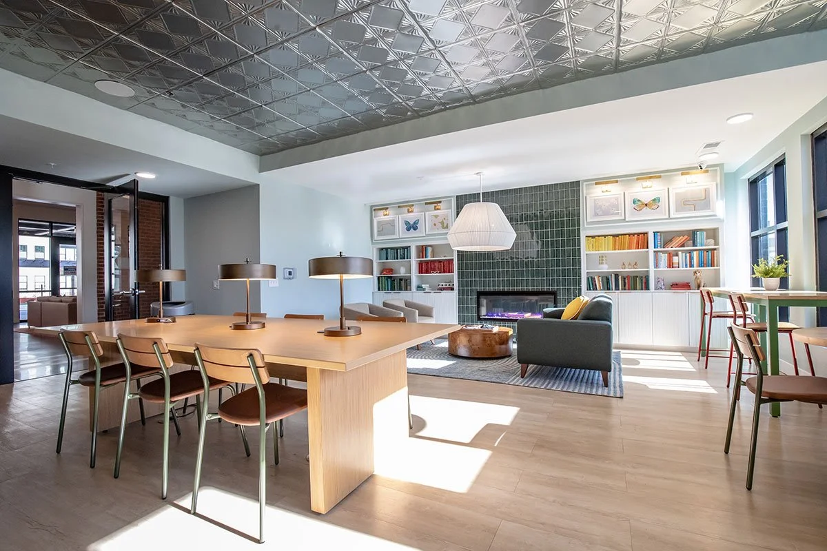 Modern living area with a wooden table, four lamps, a sitting area with a blue armchair, a green-green tiled accent wall, bookshelves, and large windows letting in natural light.