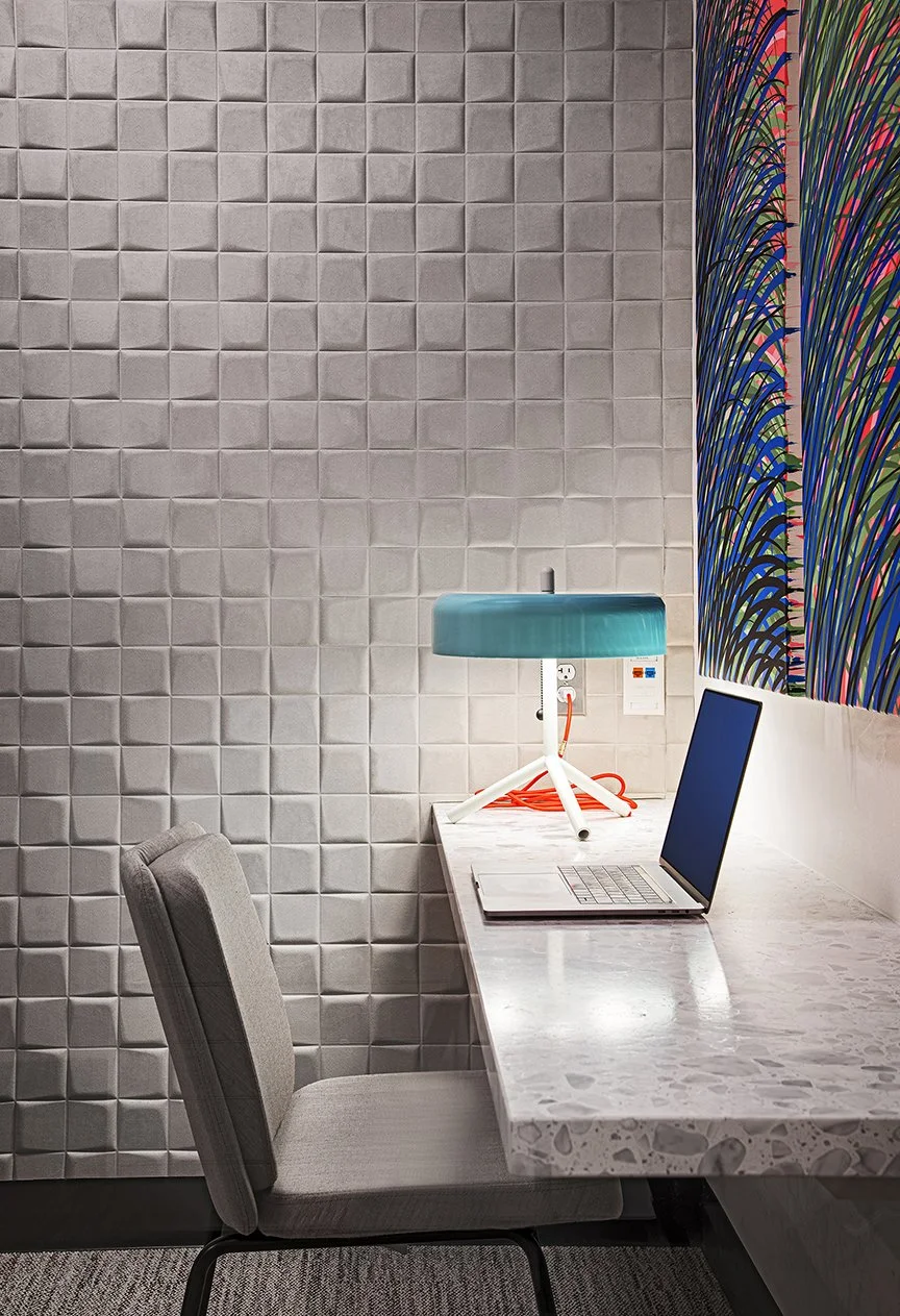 A modern workspace with a marble desk, an open laptop, a teal table lamp, a gray office chair, and a colorful abstract wall behind.