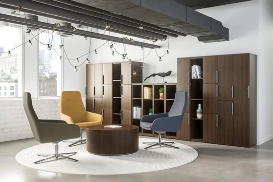 Modern office lounge with three swivel chairs in beige, blue, and gray around a round wooden coffee table, against a backdrop of bookshelf wall units, large windows, and minimalist decor, with string lights hanging from exposed ceiling beams.