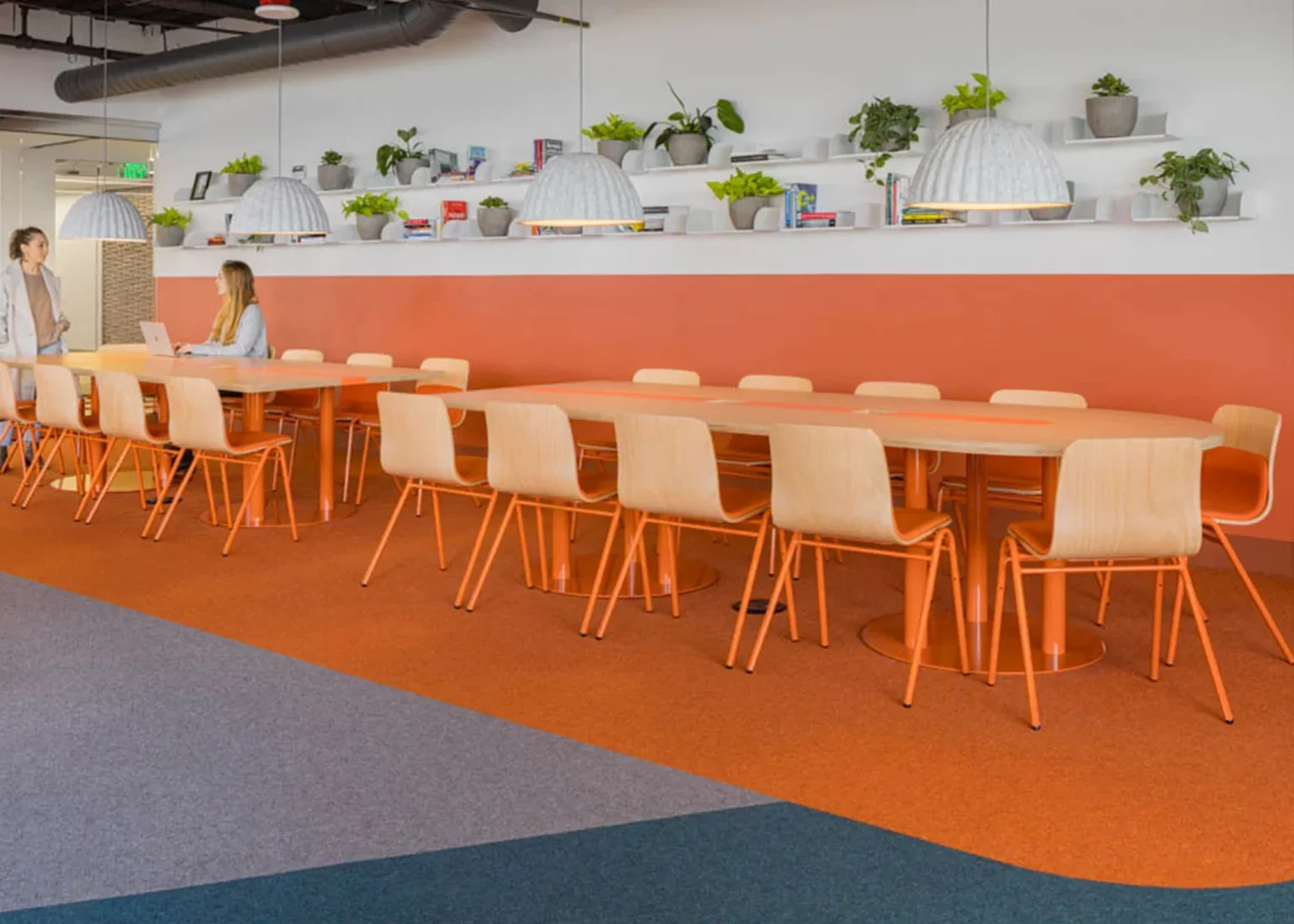 Modern office lounge with long table and beige chairs, potted plants on white shelves, two women in conversation, and a woman working on a laptop.