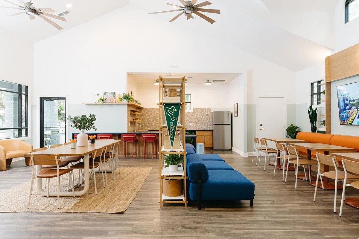 Bright, modern communal area with high vaulted ceilings, ceiling fans, large windows, a hardwood floor, a dining table with chairs, a navy blue couch, a breakfast bar with red stools, and a kitchen with stainless steel fridge.