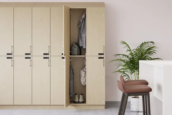 A row of beige lockers with one open, revealing hanging clothes, a backpack, and shoes inside. To the right, a potted plant, a pinkish-brown bench, and a white wall.