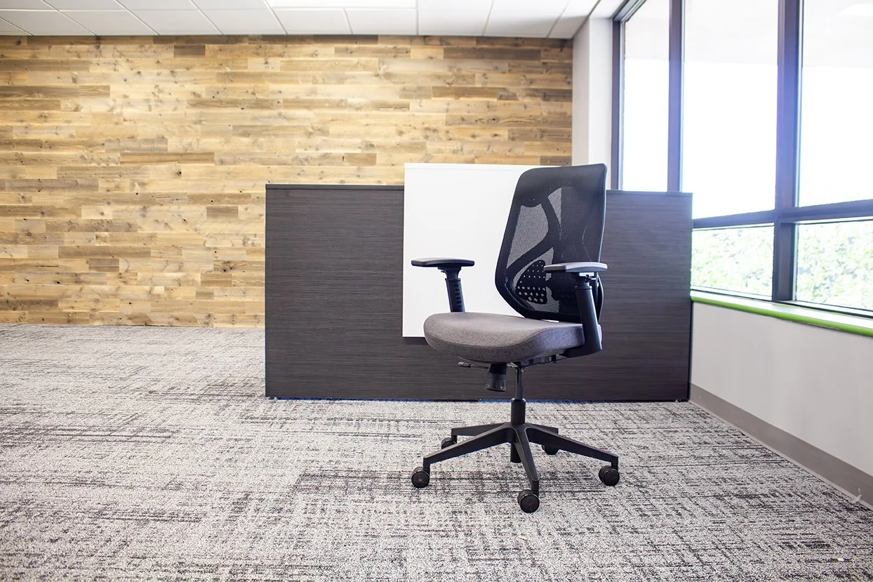 Empty office cubicle with a black mesh office chair, a wooden wall on the left, large windows on the right letting in natural light, and a carpeted floor.