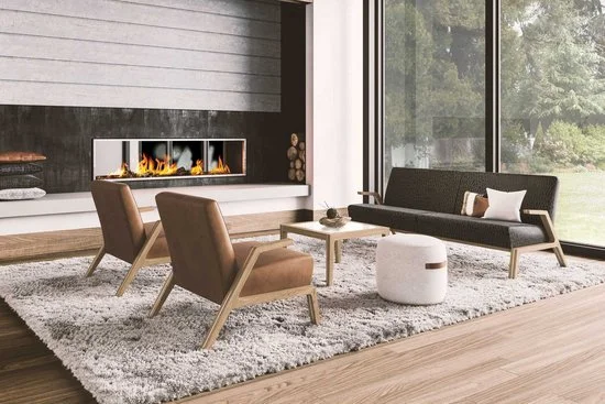 Modern living room with a fireplace, two brown chairs, a black sofa with white and colorful pillows, a white ottoman, a small table, and large windows showing greenery.